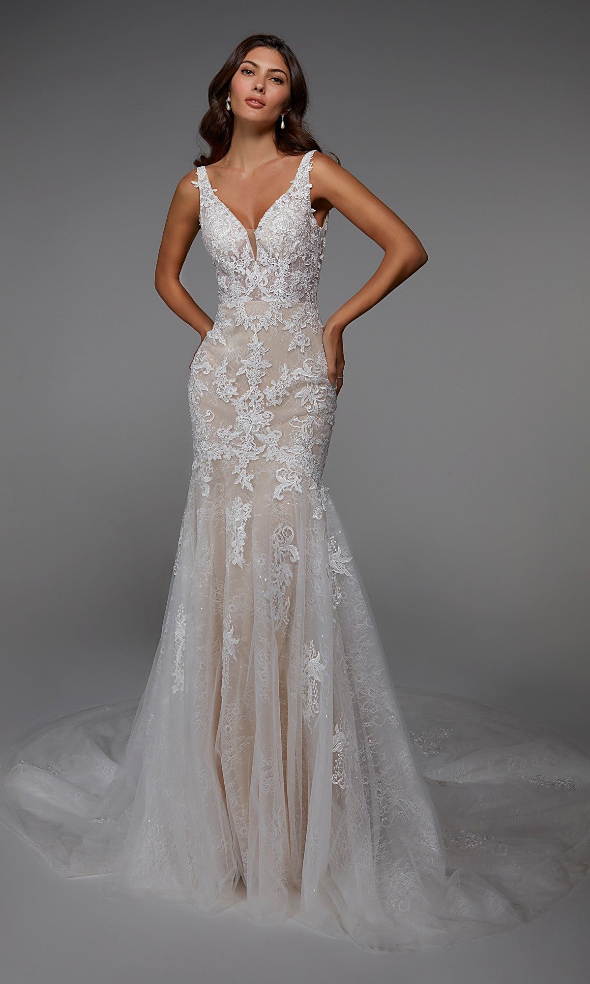 A woman poses against a plain gray background wearing the Long Formal Dress: Alyce 7029 in Ivory/Champagne, a floor-length gown with a trumpet silhouette, sleeveless design, lace details, deep V-neckline, and fitted bodice.